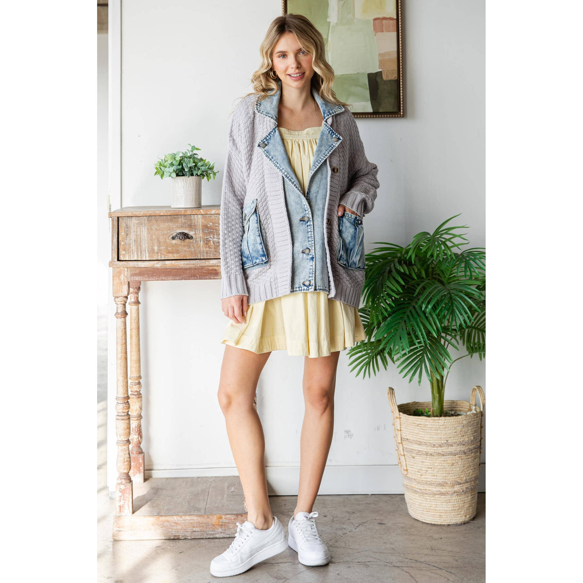 A woman with blonde hair smiles indoors by a wooden table and potted plant, wearing Veveret - Knit Body Denim Layerd Jacket: Cream / Denim / L, a yellow dress, and white sneakers.