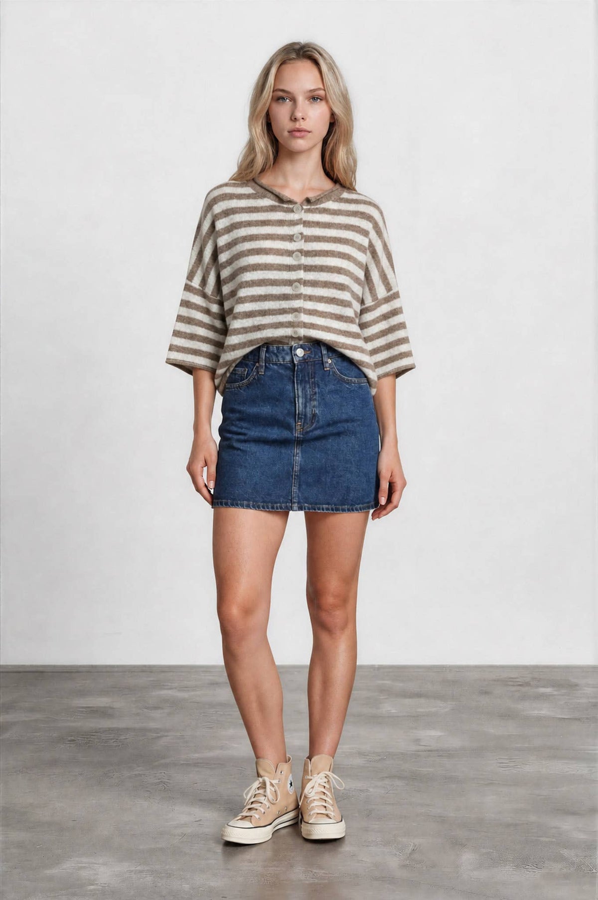 A woman stands on a concrete floor against a plain light background, wearing the Stephanie Striped Cardigan—a beige and white striped wool-blend knit sweater—paired with a blue denim skirt and beige high-top sneakers.