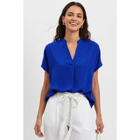 A smiling woman wears a Naked Zebra - Drop Shoulder Regular Blouse: Chocolate / L, a cobalt blue, drop shoulder woven polyester blouse with a V-neck, paired with white trousers with a drawstring waist.