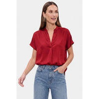 A woman wears a Naked Zebra - Drop Shoulder Regular Blouse: Chocolate / L, a short-sleeved, dark red drop shoulder blouse with a split neckline and blue jeans.