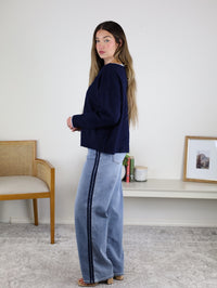 A woman with long hair wears the Bell Contrast Boat Neck Sweater and wide-leg jeans with a dark side stripe, standing indoors by a wooden chair, with a white shelf and framed photo in the background.