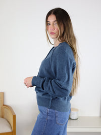 A woman with long brown hair stands sideways indoors, wearing the Theo Sweater and jeans. She looks at the camera beside a white table with a candle and a wooden chair.