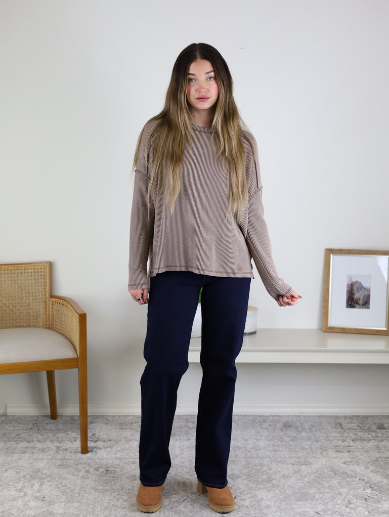 A woman with long hair indoors wears a loose beige Finley Sweater with raw edge details, dark pants, and tan shoes. She stands on a light carpet between a wooden chair and a low white shelf.
