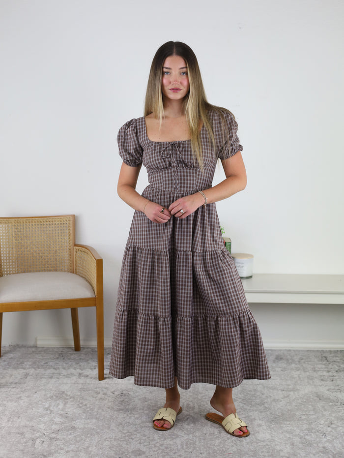 A woman stands indoors wearing the Fable Puff Sleeve Midi Dress—a brown plaid midi dress with puff sleeves and a tiered skirt—paired with beige sandals and subtle jewelry. She is positioned in front of a chair and white wall, standing on a light gray carpet.