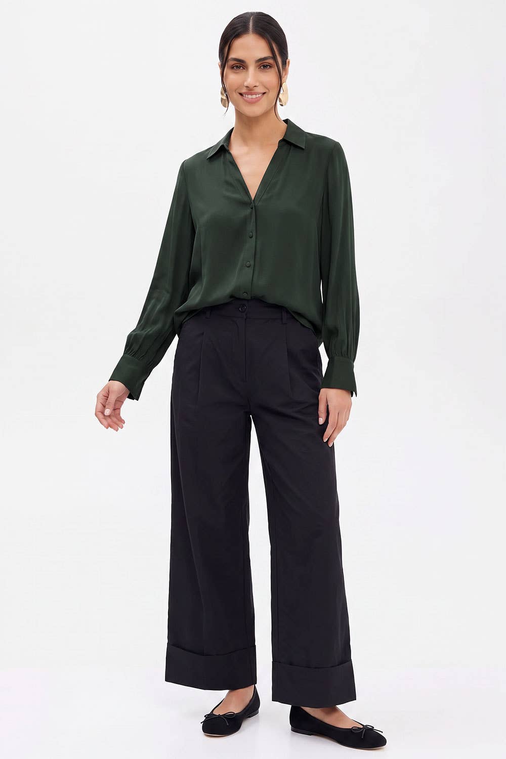 A woman wears a long-sleeved, dark green Naked Zebra - Neck Shirt With Open Seam Sleeve: Wine / L with open seam sleeves tucked into black wide-leg trousers.