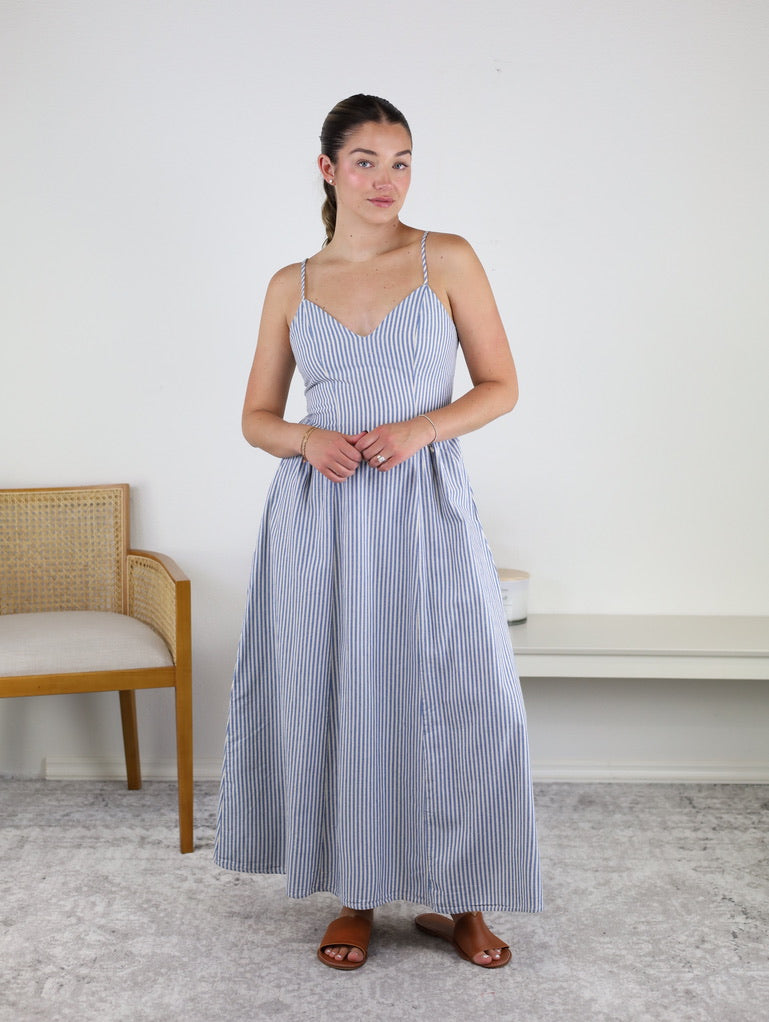 A woman stands indoors wearing the Molly Striped Maxi Dress, showcasing its blue and white stripes and adjustable straps for a perfect fit. Crafted from lightweight summer cotton, this dress is paired with brown sandals as she poses with her hands clasped in front of her. A wooden chair and white bench are visible in the background.
