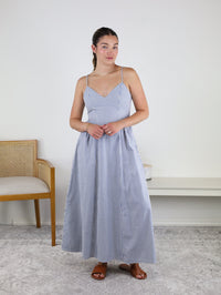 A woman stands indoors wearing the Molly Striped Maxi Dress, showcasing its blue and white stripes and adjustable straps for a perfect fit. Crafted from lightweight summer cotton, this dress is paired with brown sandals as she poses with her hands clasped in front of her. A wooden chair and white bench are visible in the background.