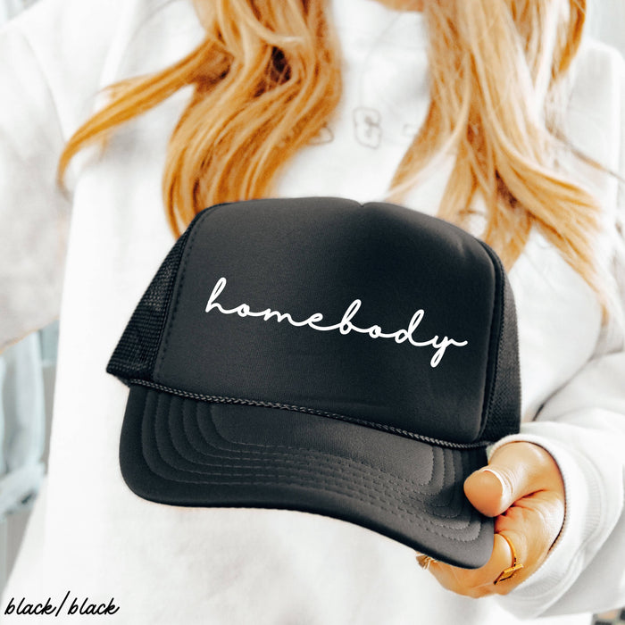 A person with long hair holds the Homebody Trucker Hat, which is black with “homebody” written in white script. The person is wearing a white sweatshirt, and the words “black/black” appear in the corner.