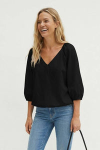 A smiling woman wears a burgundy Naked Zebra - Elastic Puff Sleeve V-neck Blouse with three-quarter length puff sleeves and blue jeans.