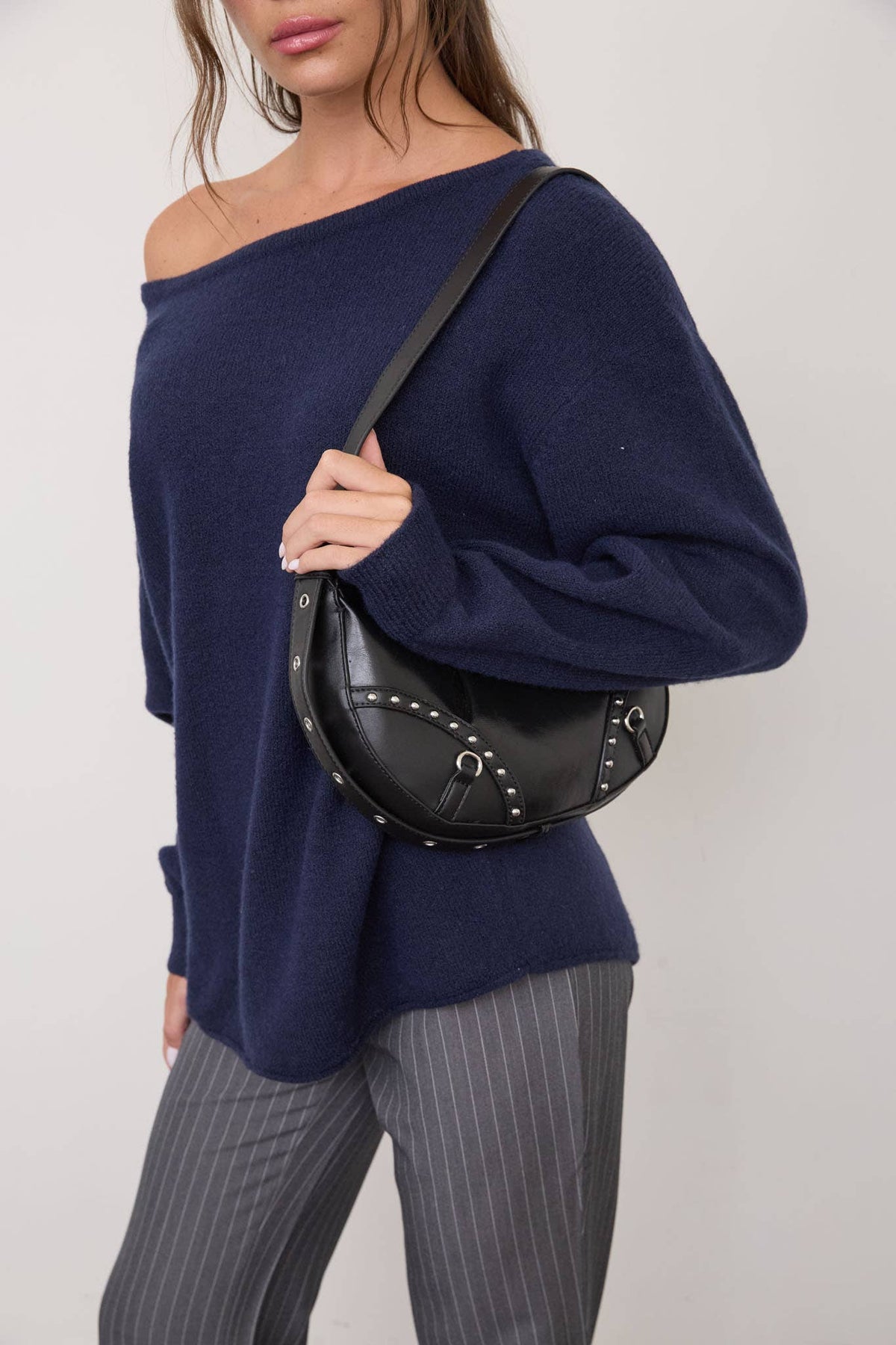 A woman wears The Workshop Studio’s Boat Neck Oversized Knit Pullover Sweater in Red Wood, styled with gray pinstripe pants and carrying a black studded handbag over her shoulder, as she stands against a plain white background.
