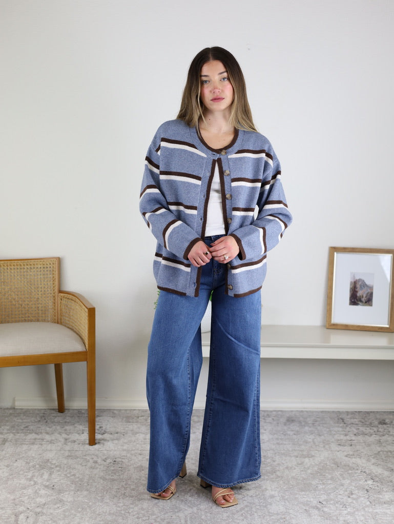 A woman wearing a blue and brown striped Nora Stripe Button Down Knit Cardigan over a white shirt and wide-leg blue jeans.