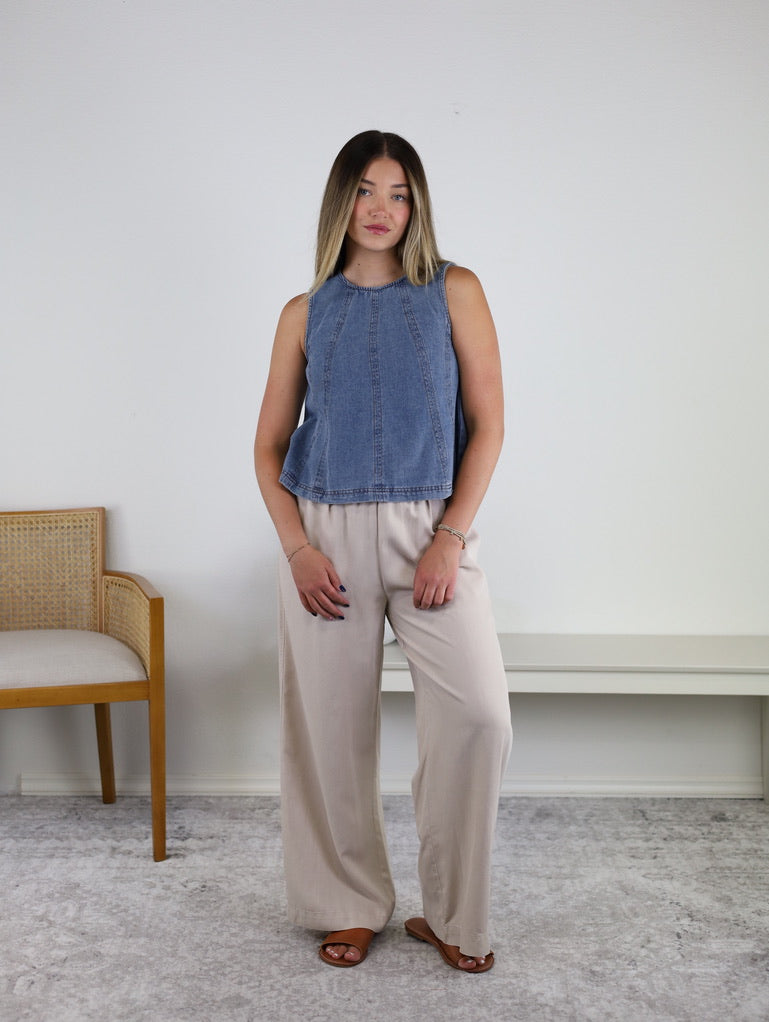 A woman stands indoors in front of a white wall, beside a wooden chair with a cushioned seat, wearing the Rebecca Seam Detail Sleeveless Denim Top – Final Sale, styled as a versatile summer blouse with beige wide-leg pants and brown sandals.