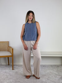 A woman stands indoors in front of a white wall, beside a wooden chair with a cushioned seat, wearing the Rebecca Seam Detail Sleeveless Denim Top – Final Sale, styled as a versatile summer blouse with beige wide-leg pants and brown sandals.