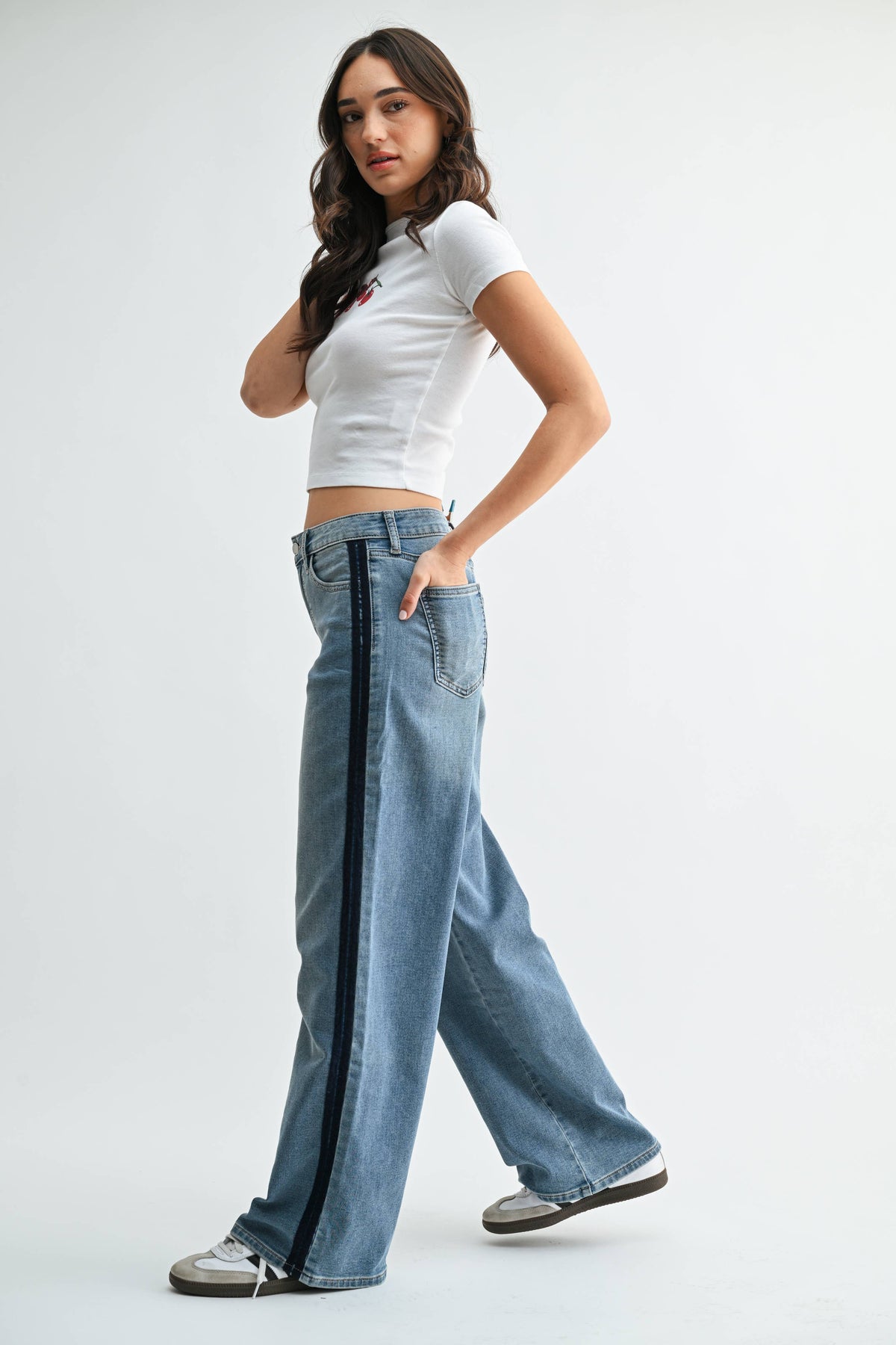 A woman stands sideways wearing a white crop top and the JBD Mid Rise Sleek Tuxedo Sweat Pants in Medium Denim, which feature a dark side stripe. With one hand in her pocket, she looks toward the camera.