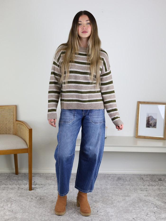 A person wears an Alexandria Striped Classic Sweater in brown, olive green, and cream, paired with loose-fit blue jeans and tan ankle boots.