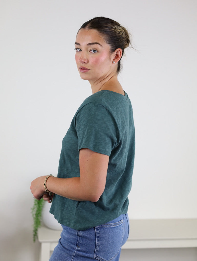 A woman with brown hair styled in a bun, dressed in the relaxed fit Nomad V Neck Tee in teal crafted from soft knit fabric and paired with blue jeans, stands sideways and looks at the camera against a plain light background.
