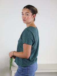 A woman with brown hair styled in a bun, dressed in the relaxed fit Nomad V Neck Tee in teal crafted from soft knit fabric and paired with blue jeans, stands sideways and looks at the camera against a plain light background.
