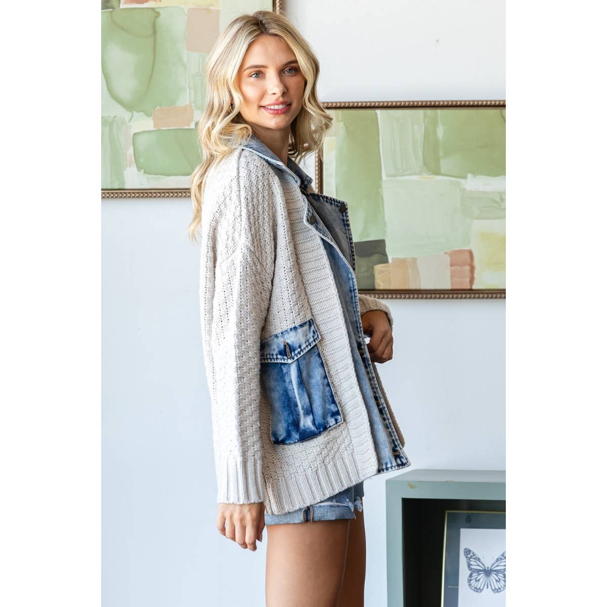 A woman with long blonde hair smiles indoors near abstract green art, wearing the Veveret - Knit Body Denim Layerd Jacket: Cream / Denim / L and denim shorts.