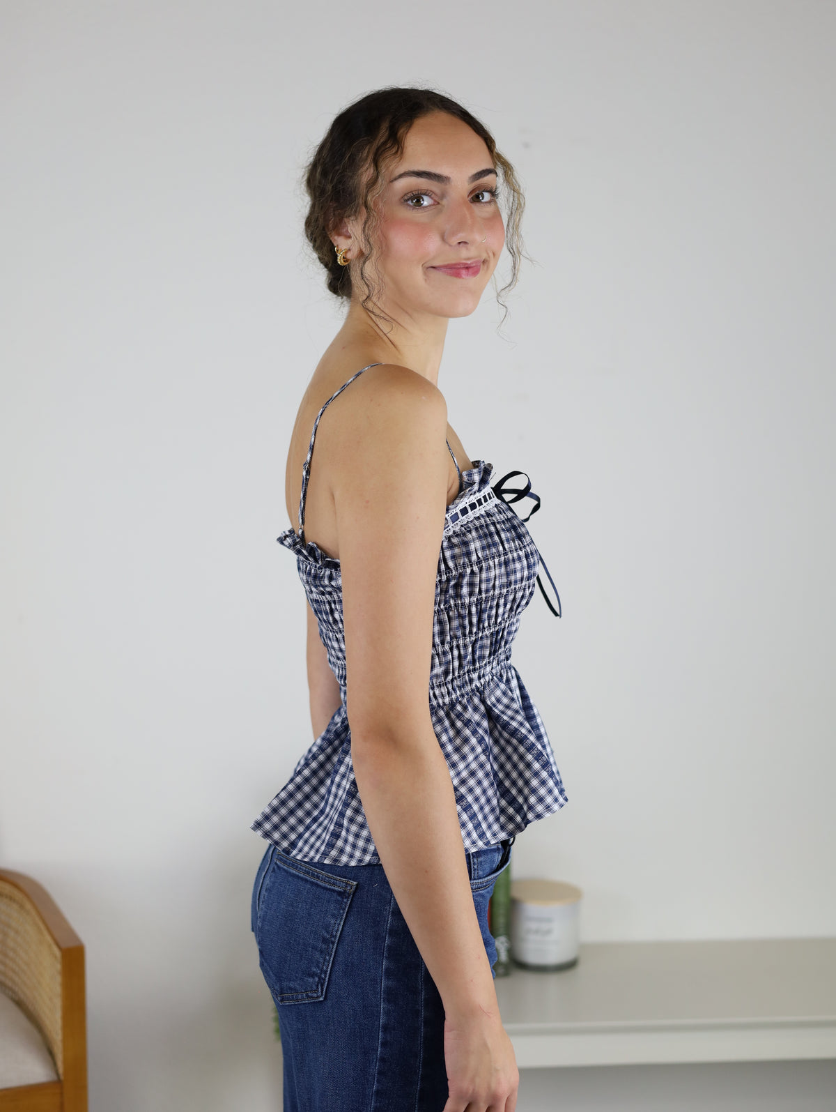 A woman with curly hair in a ponytail, wearing the Lonnie Smocked Gingham Peplum Top with adjustable spaghetti straps and blue jeans, stands sideways and smiles at the camera in a simple indoor setting.