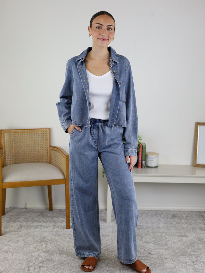 A woman stands indoors, smiling, wearing the Adalyn Acid Wash Denim Boxy Shirt paired with denim pants and a white top. Behind her are a wooden chair, a shelf with plants, a candle, a framed mirror, and a white rug on the floor.