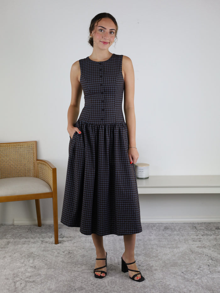 A woman stands indoors wearing the Vivi Round Neck Button Front Midi Dress, styled with black-heeled sandals. She is positioned near a bench and a wooden chair against a plain white wall.