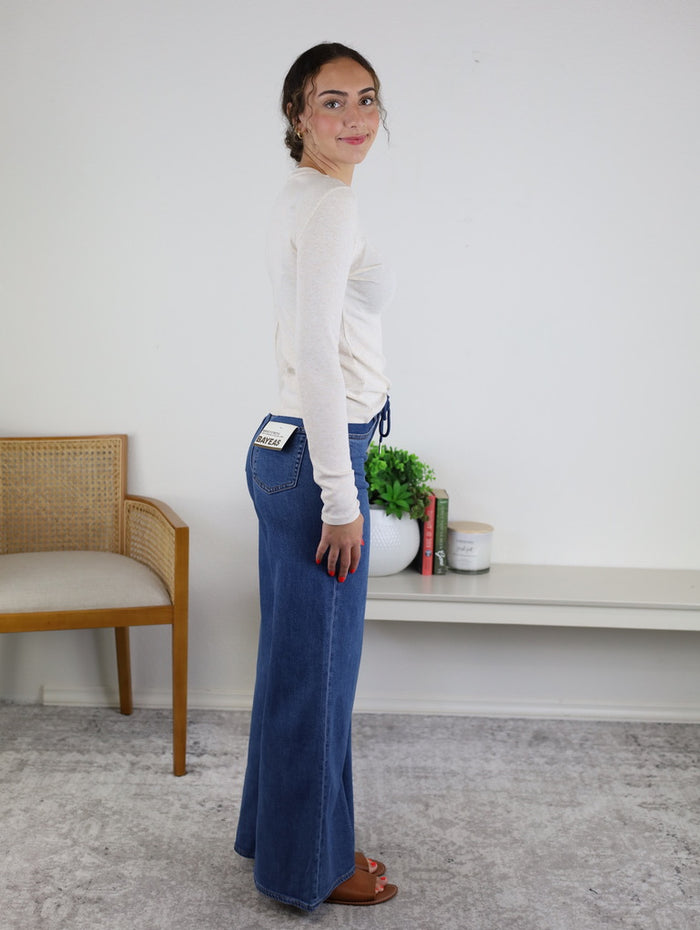 A person with curly hair stands sideways, wearing a long-sleeve white top, the Rain Mid Rise Wide Leg jeans, and sandals. There is a chair, plant, books, and a candle on a bench in the background.