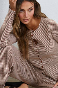A woman with long wavy hair wears the Creamon - CTP3592 BROWN Lounge Striped Button Down Top & Pants Set, featuring brown and white stripes with a button down top and matching wide leg pants. She sits with one arm resting on her knee, looking to the side.
