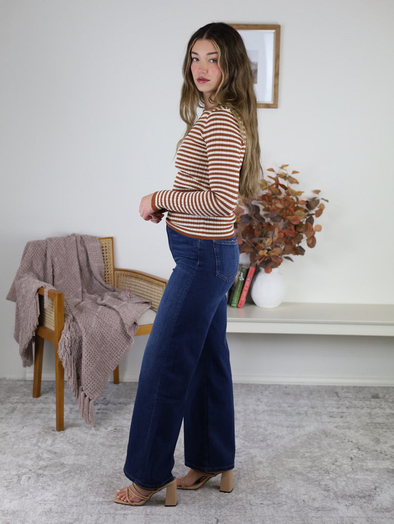 A woman wearing a terracotta stripe ribbed knit Mindy Ribbed Striped Top with long sleeves, paired with dark wash wide-leg jeans and tan heeled sandals.