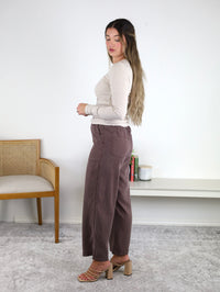 A woman with long hair, dressed in a beige top and brown Marbella Wide Leg Tencel Pant, stands sideways in a minimal room featuring a wooden chair, white bench, and several books in the background. She wears heeled sandals and looks downward, highlighting the breathable, flowy fit and comfortable drawstring elastic waist of the Marbella Wide Leg Tencel Pant for an effortlessly stylish look.