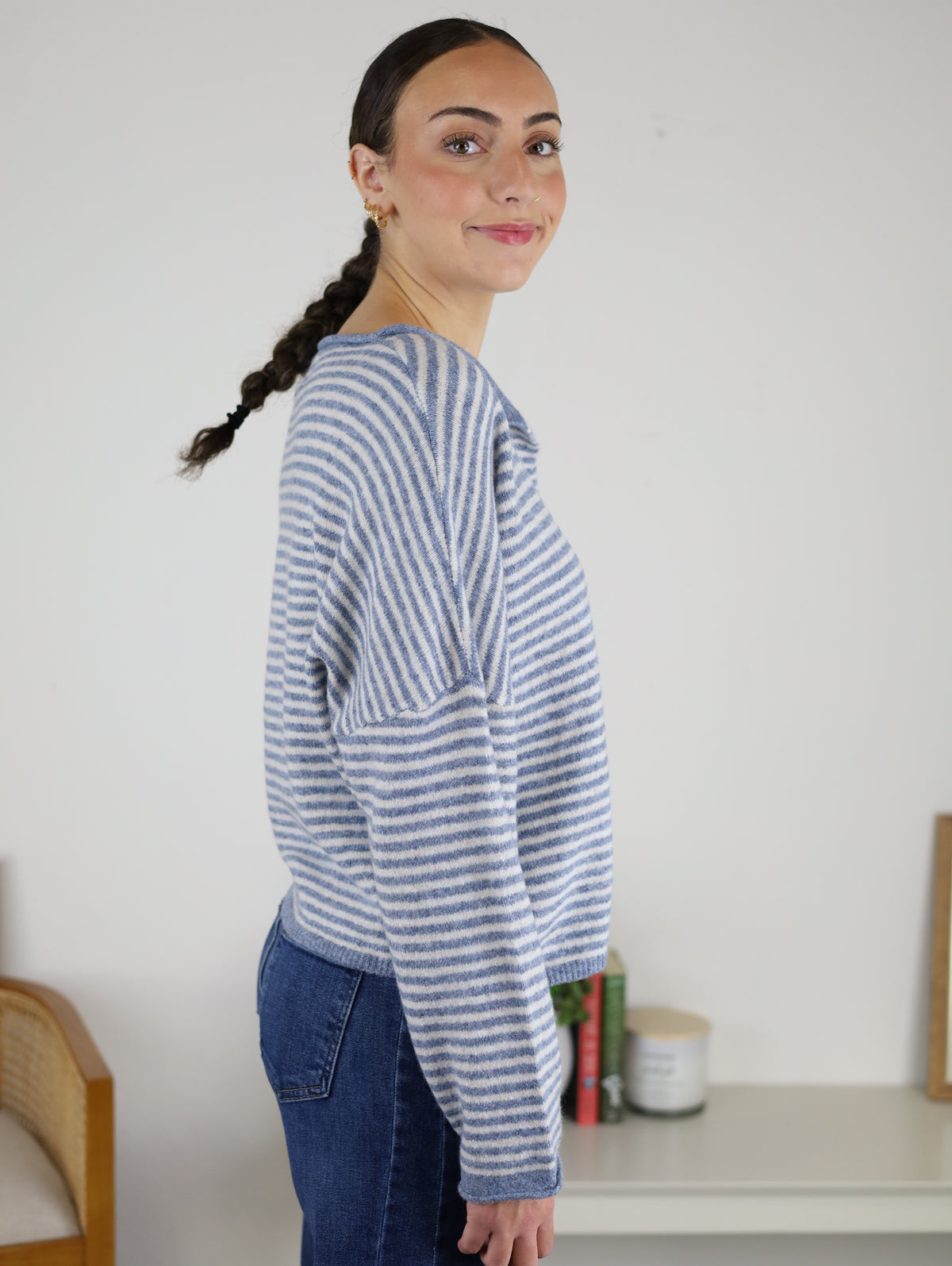 A woman with braided hair stands sideways and smiles, wearing the Maren Striped Cardigan in a relaxed fit paired with jeans. In the background, a white shelf displays books and a candle.