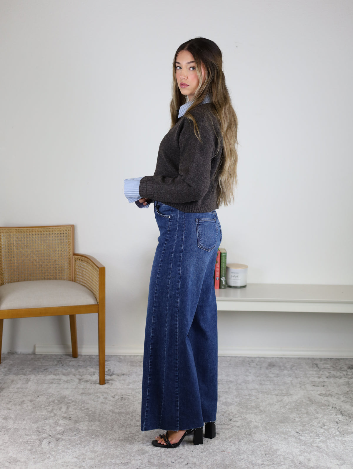 A woman with long hair stands sideways in a room, wearing the Mani Stripe Trim Button Down Cardigan over blue wide-leg jeans and black heels. A wooden chair and a low shelf with books are behind her.