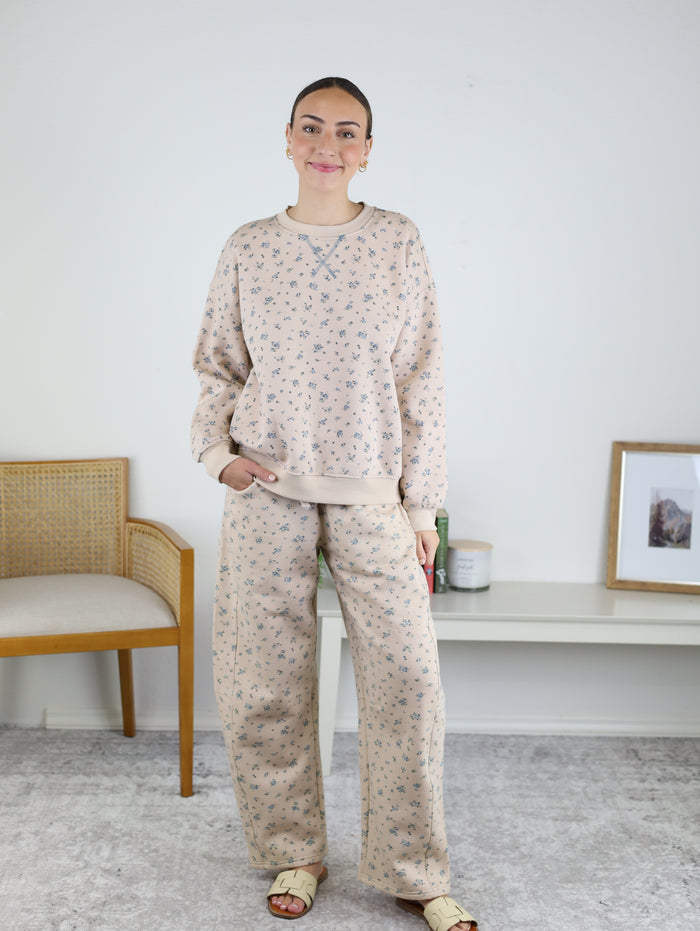 A woman stands indoors, smiling with one hand in her pocket, wearing the Journey Floral Boxy Crewneck Lounge Set, which features a khaki floral crewneck sweatshirt and wide-leg pants. A chair, table, and framed picture are visible in the background.