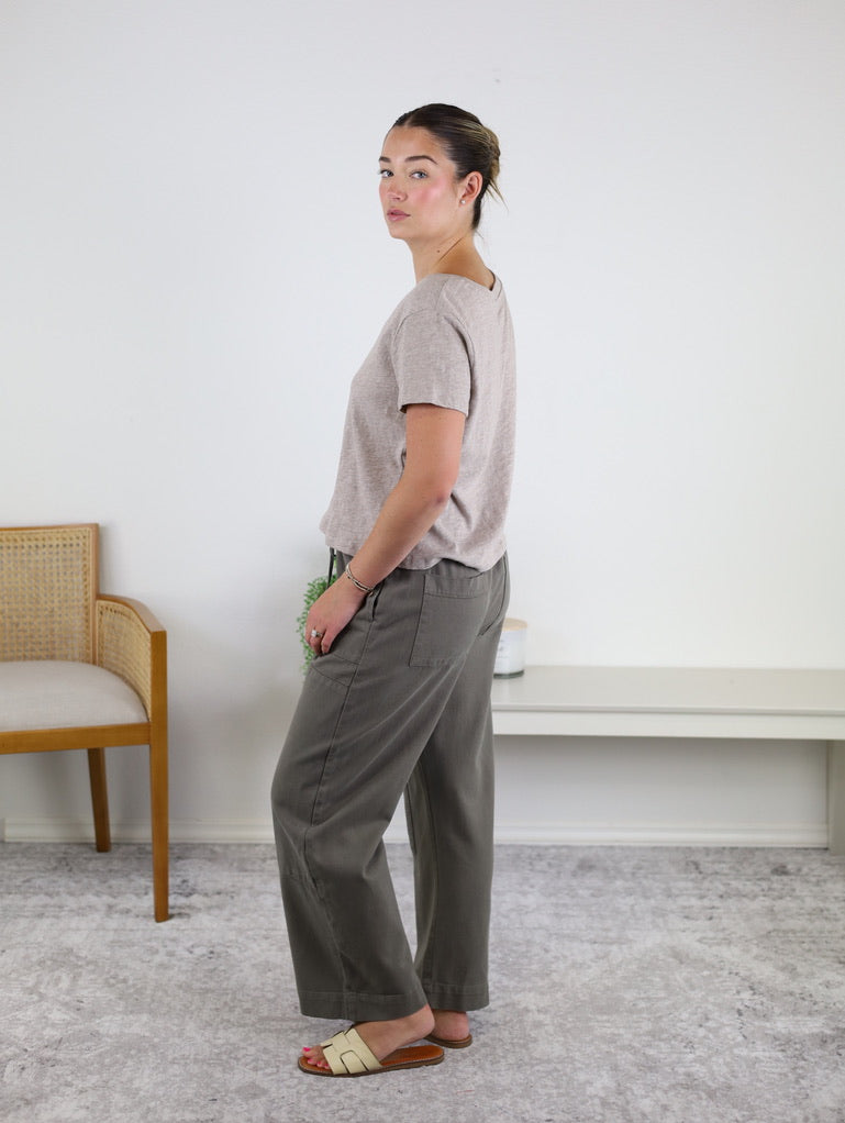 A woman stands sideways in a light room, wearing the Nomad V Neck Tee in a relaxed fit beige knit, paired with grayish-green pants and white sandals. In the background, there is a wooden chair and a white bench with a small plant.