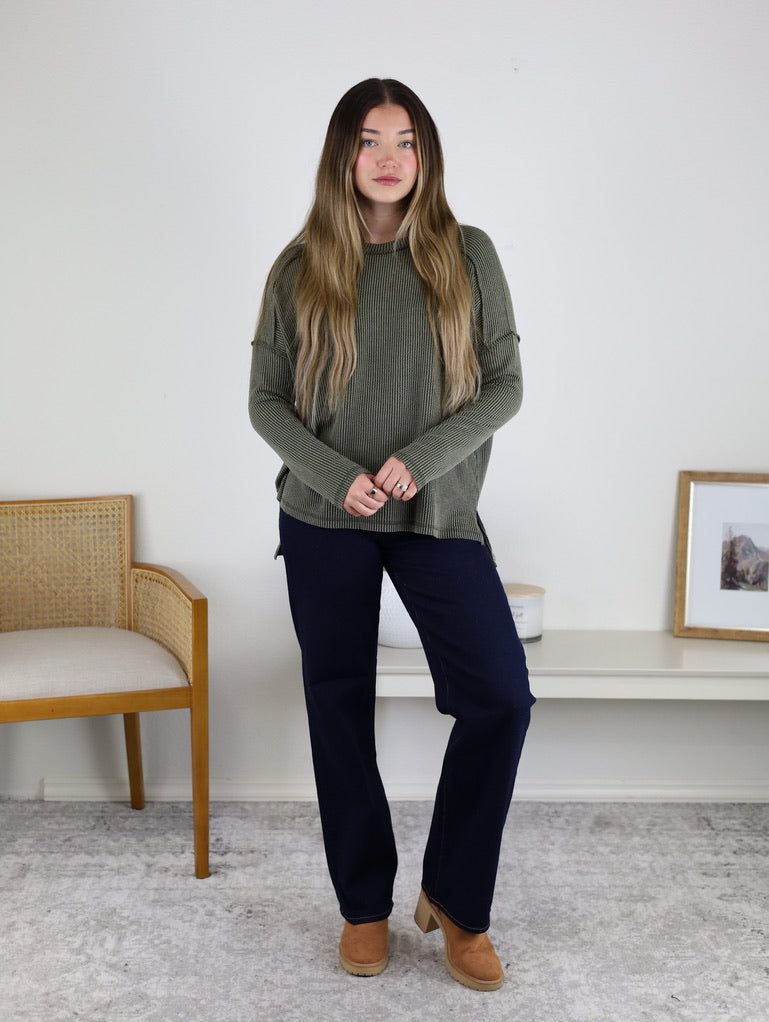 A woman with long hair stands indoors wearing the Finley Sweater in green, dark blue pants, and tan shoes. She is in front of a white wall, with a chair to her left and art on a low table to her right.