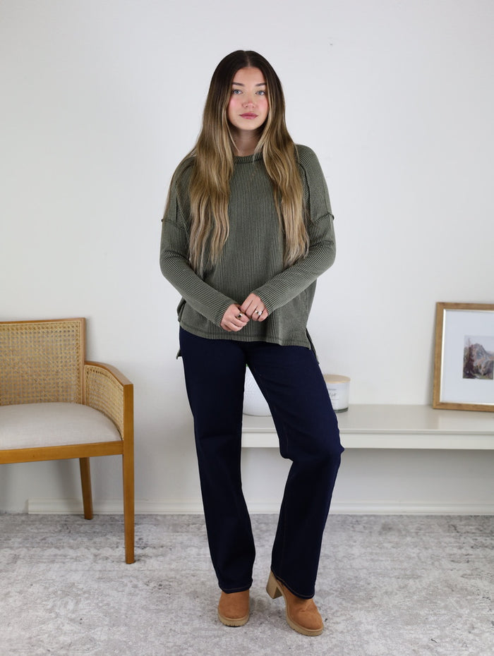 A woman with long hair stands indoors wearing the Finley Sweater in green, dark blue pants, and tan shoes. She is in front of a white wall, with a chair to her left and art on a low table to her right.