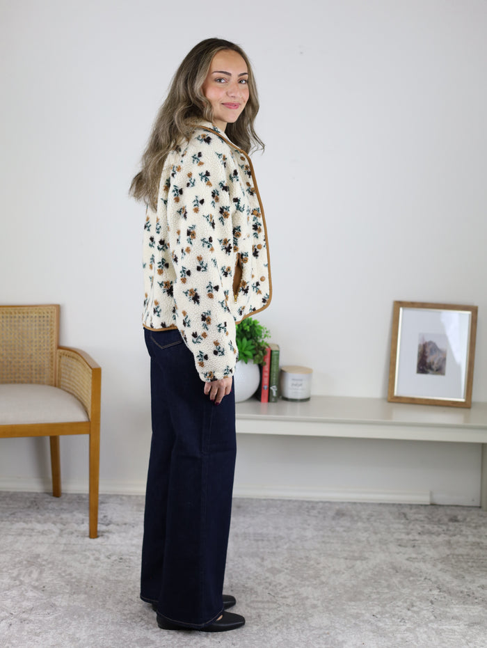 A woman wears a cozy, cream-colored Casin Floral Print Fluffy Fleece Jacket with brown trim and dark blue wide-leg jeans.