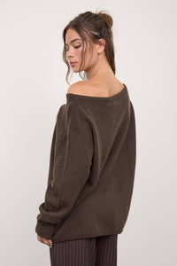 Wearing The Workshop Studio’s Boat Neck Oversized Knit Pullover Sweater in Red Wood, a woman with loosely tied hair looks down while standing against a plain light background, paired with pinstriped pants.