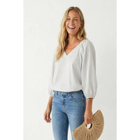 A smiling woman wears a burgundy Naked Zebra - Elastic Puff Sleeve V-neck Blouse with elastic puff sleeves and blue jeans, holding a crescent-shaped straw handbag.