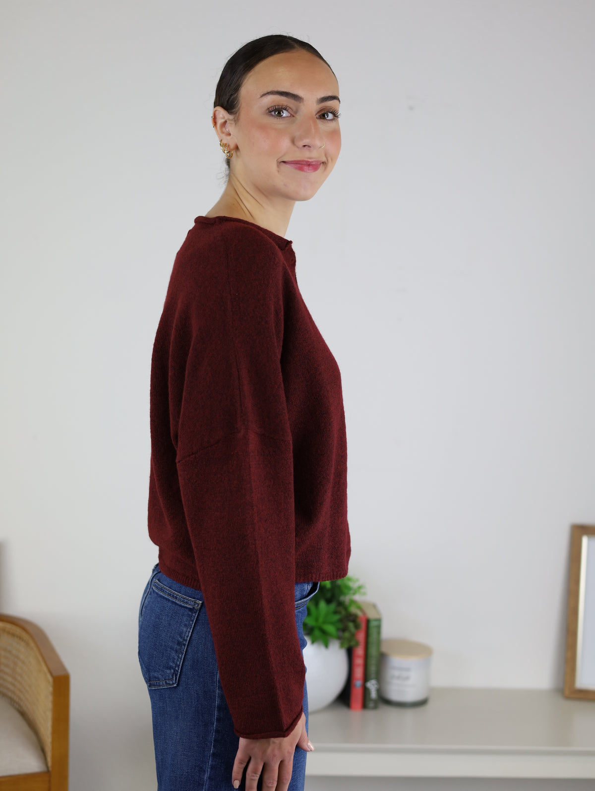 A woman with dark hair in a bun, wearing the Beth Lightweight Cardigan in burgundy and blue jeans, stands sideways and smiles. Behind her are books, a potted plant, and a candle on a white table against a plain wall.