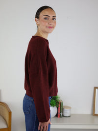 A woman with dark hair in a bun, wearing the Beth Lightweight Cardigan in burgundy and blue jeans, stands sideways and smiles. Behind her are books, a potted plant, and a candle on a white table against a plain wall.