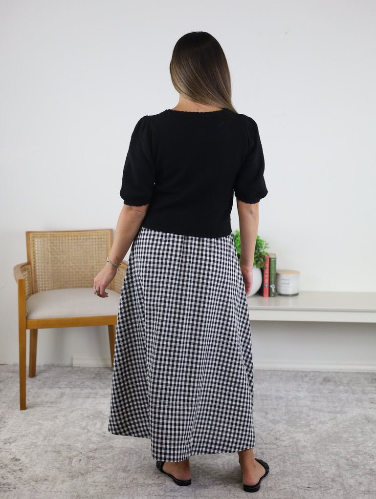 A woman stands indoors with her back to the camera, barefoot on a light rug, wearing the Monica Short Sleeve Cardigan in black, featuring metallic buttons, paired with a black-and-white checkered skirt. A wooden chair and a white shelf are visible in the background.