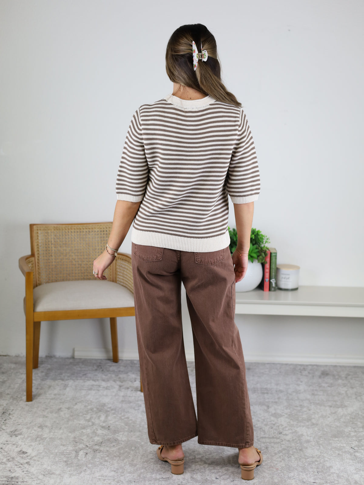 A person stands facing away, wearing the Scout Striped Sweater, brown wide-leg pants, and sandals. Their hair is clipped back. A chair, shelf with books, and plants are visible in the background.