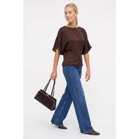 A person walking in a Naked Zebra - Back Tie Dolman Sleeve Silky Fall Blouse: Wine / L, paired with blue wide-leg jeans and pointed-toe shoes, carrying a brown structured handbag.