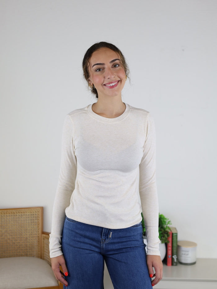 A woman with curly hair wearing the Josette Ribbed Classic Crewneck and blue jeans smiles while standing indoors near a chair and a small table with books and a potted plant.