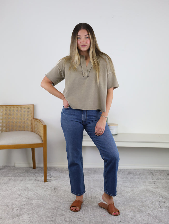 A person stands indoors wearing the Wilder Split Neck Top in beige, featuring a subtle washed finish, paired with blue jeans and brown sandals. Their left hand rests on their hip, with a wooden chair and a white bench visible in the background.