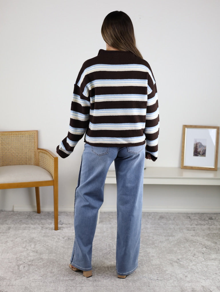 A person seen from behind, wearing blue jeans and a Remy Multicolor Rolled Neck Knit Cardigan featuring blue, brown, and white stripes with a rolled neck.