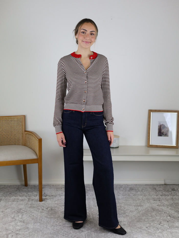 A person wears a Vera Striped Cardigan in brown and white horizontal stripes with red trim and buttons, paired with dark wash wide-leg jeans.