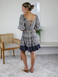 A woman wears a white and blue print Leslie Smocked Mini Dress featuring puff sleeves and a lace-up detail.