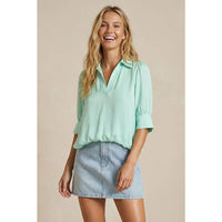 A smiling woman wears a mint green Naked Zebra - Half Sleeve Hi-lo Bubble Hem Blouse with a collared V-neck, paired with a light-wash denim mini skirt.