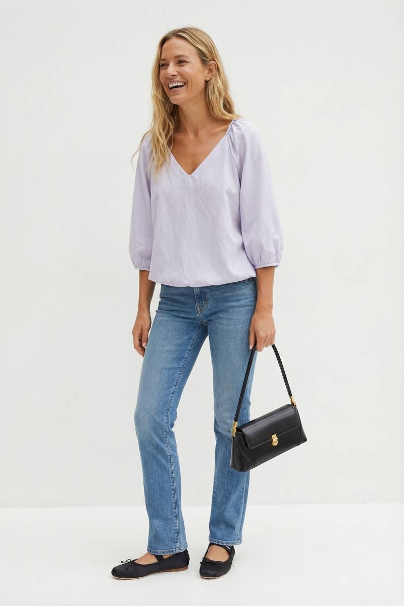 A smiling woman wearing a Naked Zebra - Elastic Puff Sleeve V-neck Blouse: Burgundy / L, blue jeans, and black ballet flats, carrying a black shoulder bag.